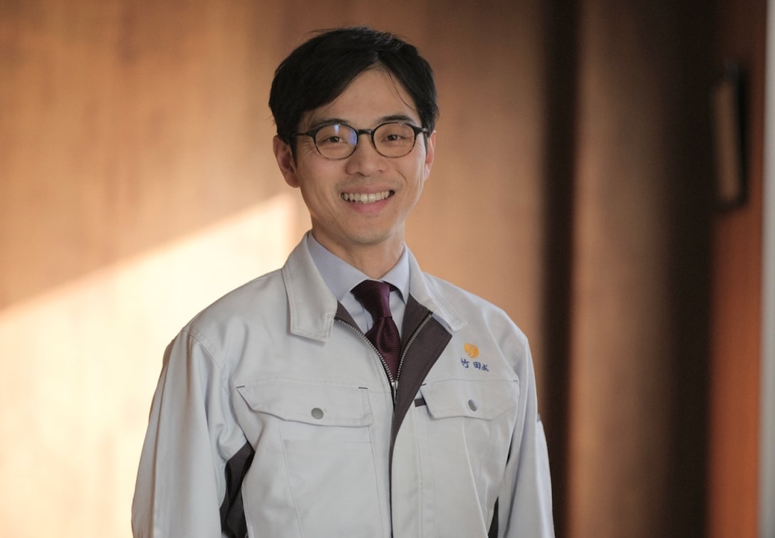 代表取締役社長 竹田成宏の写真。眼鏡をかけ、ユニフォームを着用し、笑顔を浮かべている。背景は木目調の部屋の壁が、柔らかな窓からの日差しに照らされている
