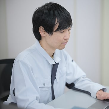 生産技術部 Sさんの写真。ユニフォームを着用し、デスクでノートパソコンに向かって作業している