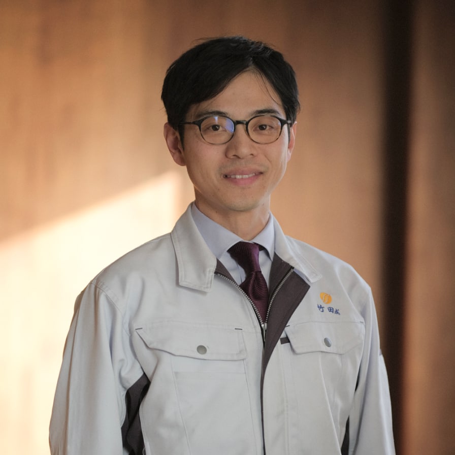 代表取締役社長 竹田成宏の写真。眼鏡をかけ、ユニフォームを着用し、微笑んでいる。背景は木目調の部屋の壁が、柔らかな窓からの日差しに照らされている