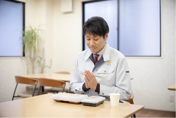 社内の休憩スペースで昼食を取る男性社員の写真。部屋の奥には観葉植物が飾られ、清潔感のある明るいスペースが広がっている