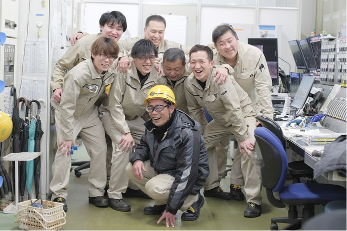 明るい笑顔を浮かべ、肩を組む8人の男性社員たちの集合写真。全員が作業着を着用し、先頭の社員はヘルメットを着用している