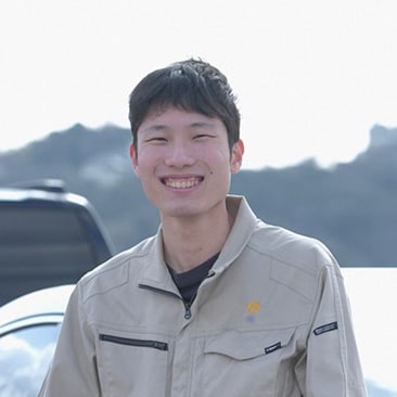 製造部 Mさんの写真。ユニフォームを着用し、笑顔を浮かべている。背景には愛車が映り込む