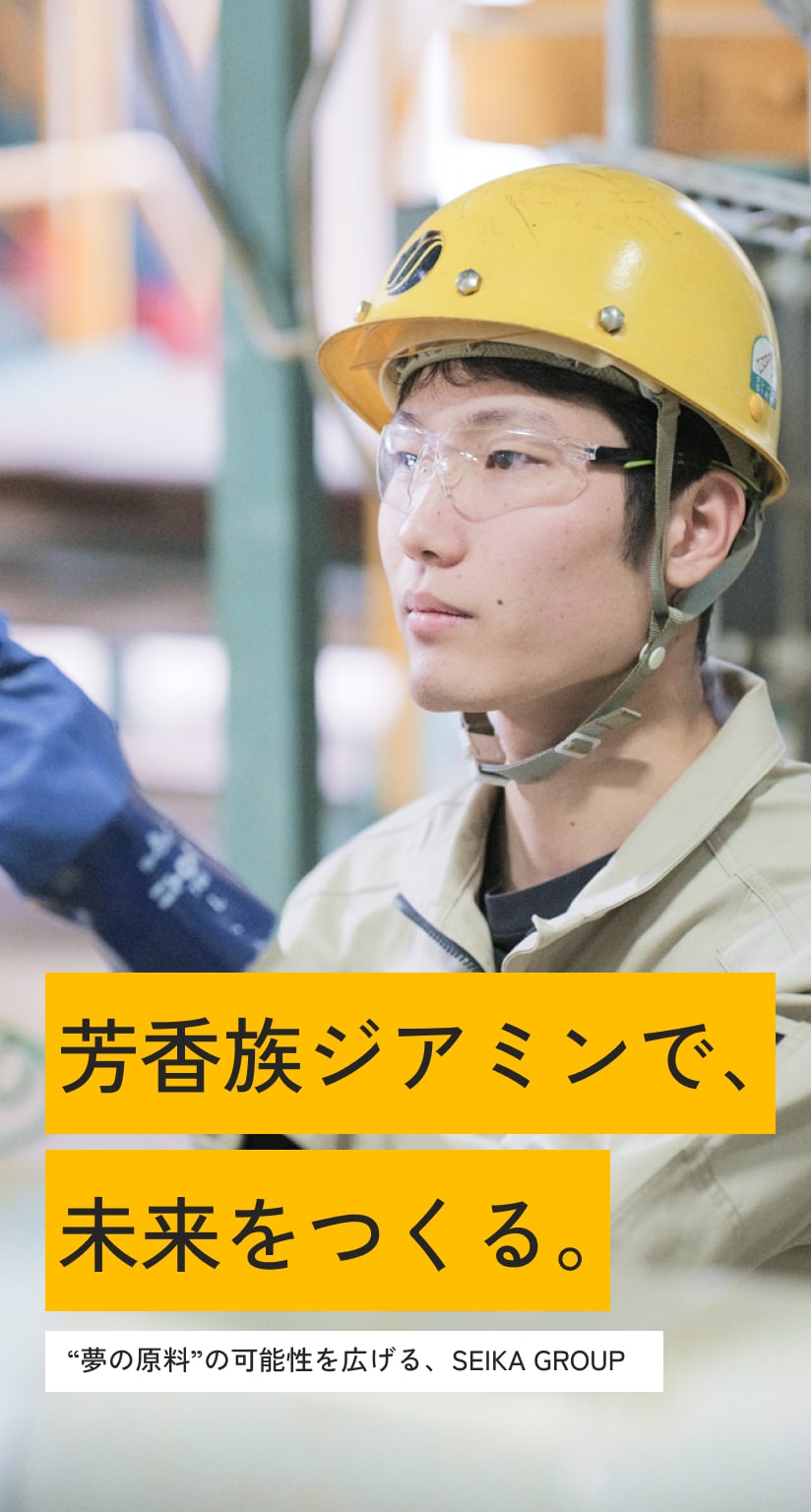 工場の機械を指差しで確認する男性社員の写真。ヘルメットと安全ゴーグル、ゴム手袋、作業着を着用している