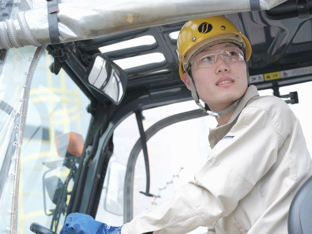 フォークリフトを運転するK.Mさんの写真。ヘルメットと安全ゴーグル、ゴム手袋、作業着を着用している