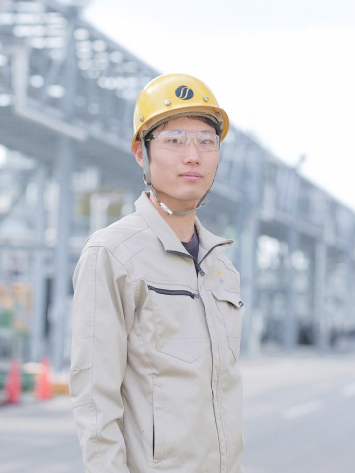 2023年度入社 製造部 K.Mさんの写真。ヘルメットと安全ゴーグルとユニフォームを着用している。背景には工場の機械や配管が映り込む