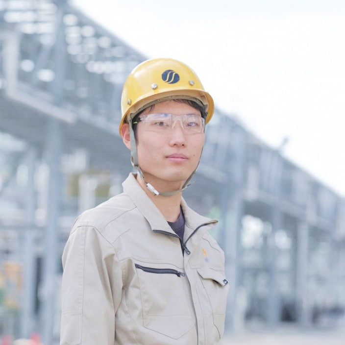 2023年度入社 製造部 K.Mさんの写真。ヘルメットと安全ゴーグルとユニフォームを着用している。背景には工場の機械や配管が映り込む
