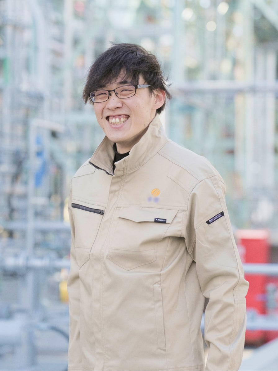 2017年度入社 製造部 S.Yさんの写真。ユニフォームを着用している。背景には工場の配管が映り込む