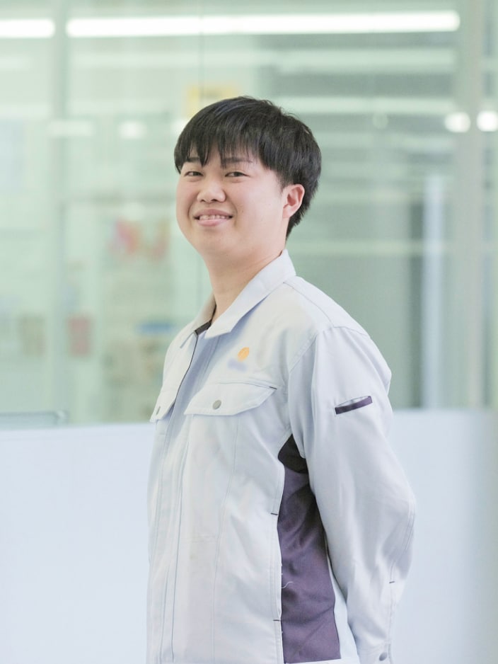2022年度入社 研究部 T.Nさんの写真。ユニフォームを着用している。背景には実験器具が並んだ部屋が映り込む