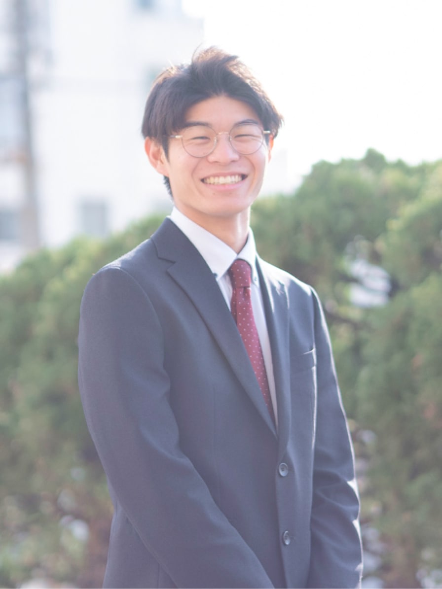 2024年度入社 営業部 T.Tさんの写真。ビジネススーツを着用している。背景には工場の遠景と大きく茂った木の緑が映り込む