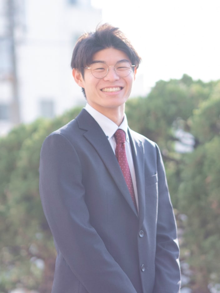 2024年度入社 営業部 T.Tさんの写真。ビジネススーツを着用している。背景には工場の遠景と大きく茂った木の緑が映り込む