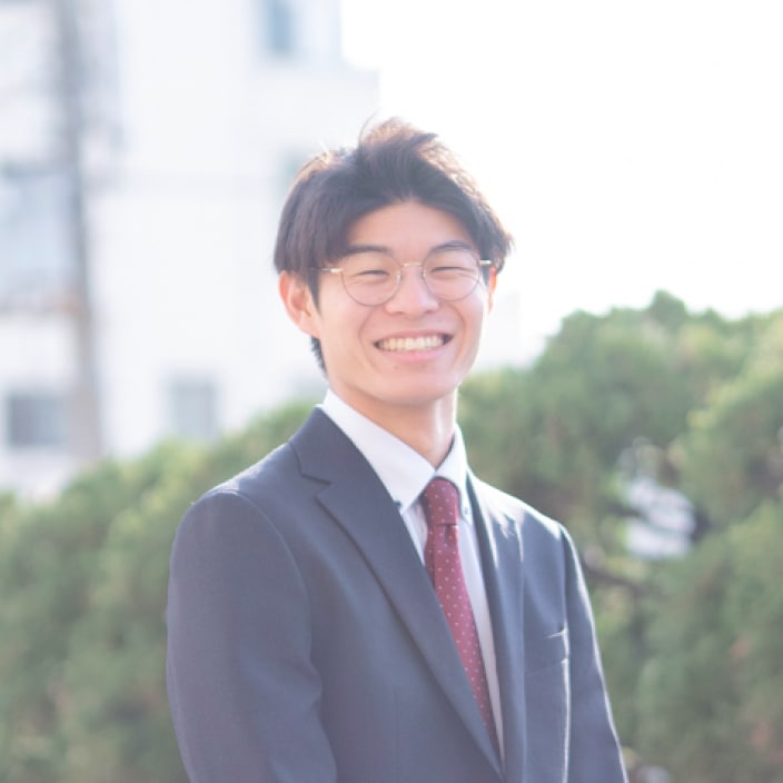 2024年度入社 営業部 T.Tさんの写真。ビジネススーツを着用している。背景には工場の遠景と大きく茂った木の緑が映り込む